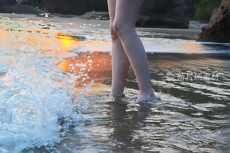 难以辨认的白人妇女在海水中划桨，海浪破浪，腿和光脚在浅滩，日落时的海边沙滩，聚焦于前景