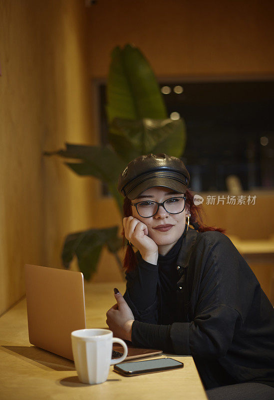 一位戴着黑色皮帽、戴着眼镜、红头发编成辫子的年轻女子，在咖啡馆里一边喝着咖啡，一边独自在电脑前工作。