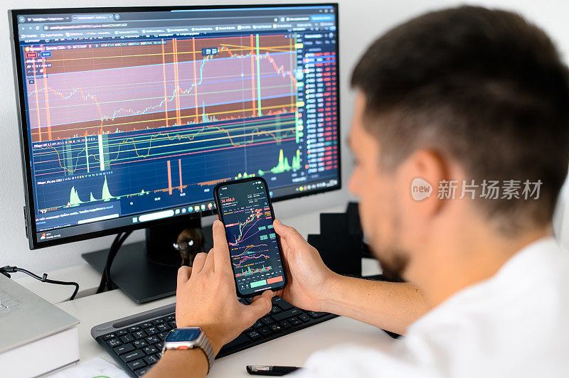 加密货币交易员坐在工作场所的显示器前，手里拿着智能手机，屏幕上显示交易数据指数图表，手机应用程序分析加密货币市场