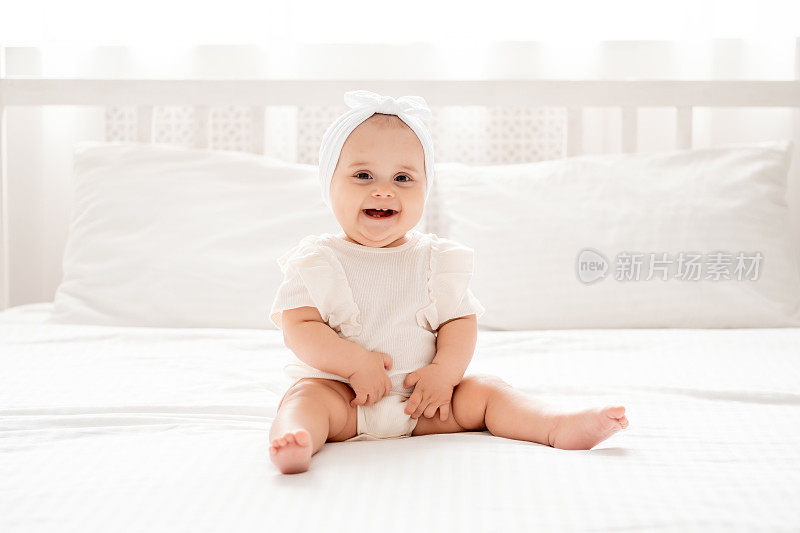 刚出生的女婴笑嘻嘻地坐在床上穿着洁白的衣服，在明亮的房间里，一个地方的文字，快乐健康的宝宝特写，孩子们的笑声