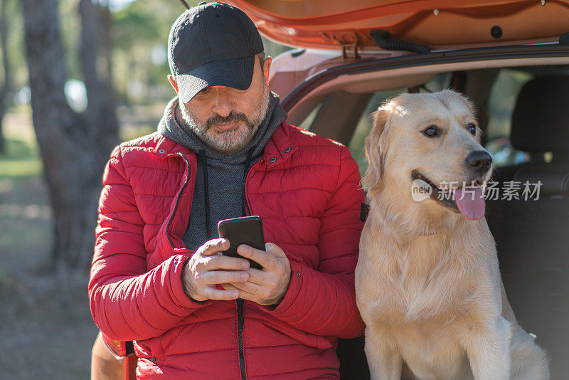 一个成熟的男人和他的狗坐在汽车后备箱上，用着智能手机