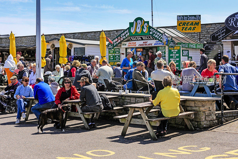 在英国多塞特的西湾，人们坐在野餐桌旁吃快餐
