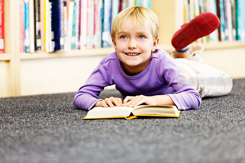 一个金发小女孩在图书馆的地板上看书，她微笑着抬起头来