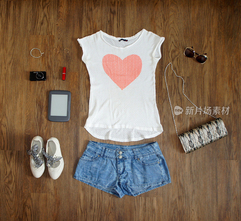 平躺女性服装和配饰拼贴:白色心形t恤、牛仔短裤、鞋子、手包、眼镜、相机、电子书、口红和木制背景上的珠宝