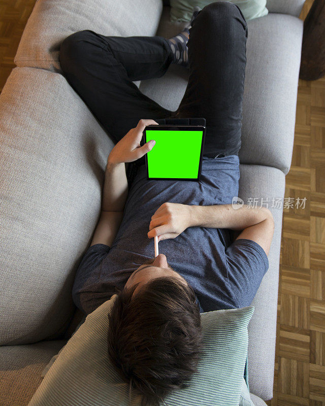 一名年轻人正在家里的沙发上用平板电脑放松