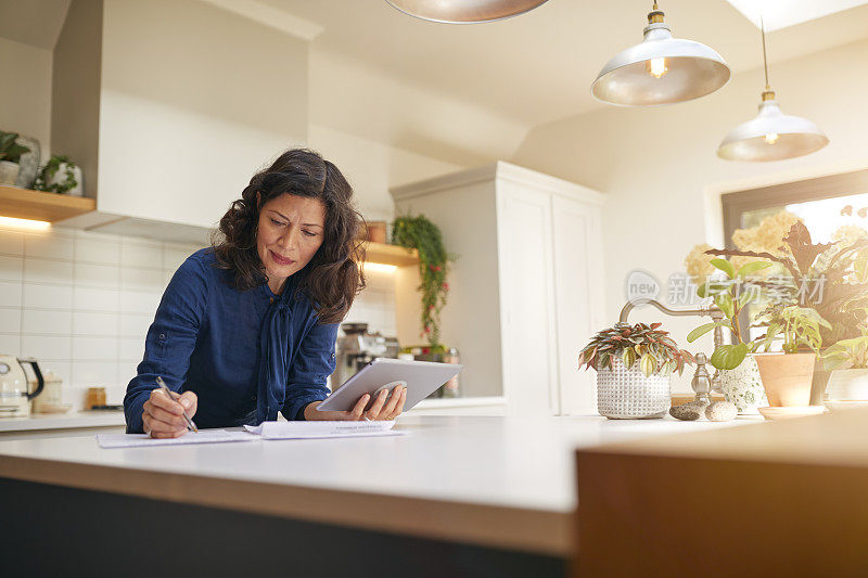 成熟的女人在家里的厨房里用平板电脑审查家庭财务和文书工作