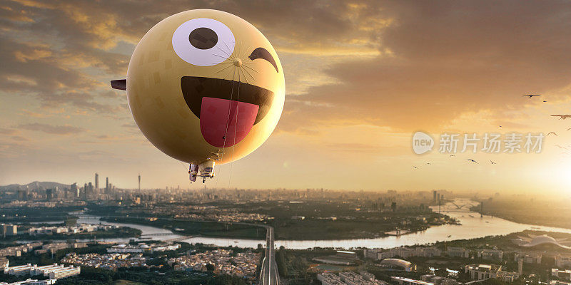 在黎明的天空中，一个带有微笑表情的球形黄色飞艇或小飞艇在城市上空飞行