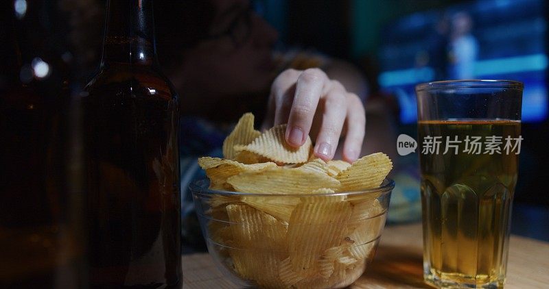 这个人正在看电视上的体育比赛，在家里喝啤酒吃薯片