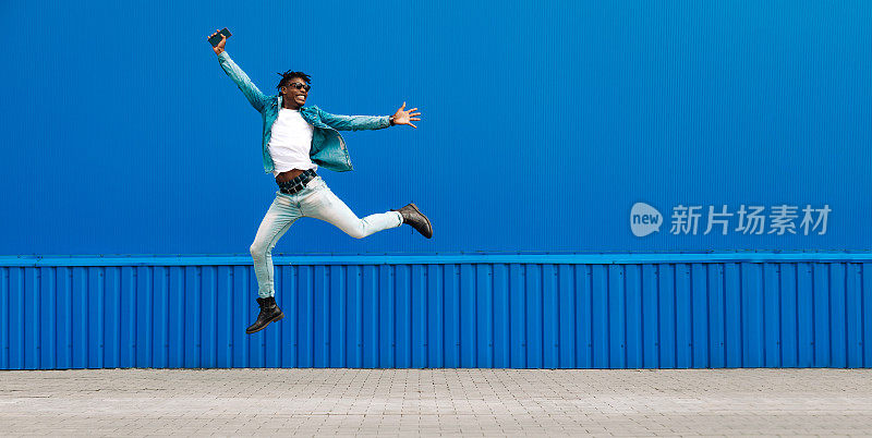 一个非洲人拿着电话跳了起来，在城市的蓝色建筑的背景下