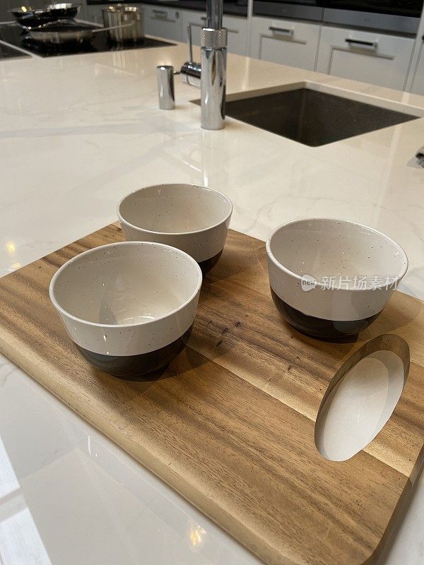 近景现代厨房设计，中央岛早餐吧，白色复合可丽安台面和木制砧板上的麦片碗，橱柜，水槽和搅拌机龙头背景