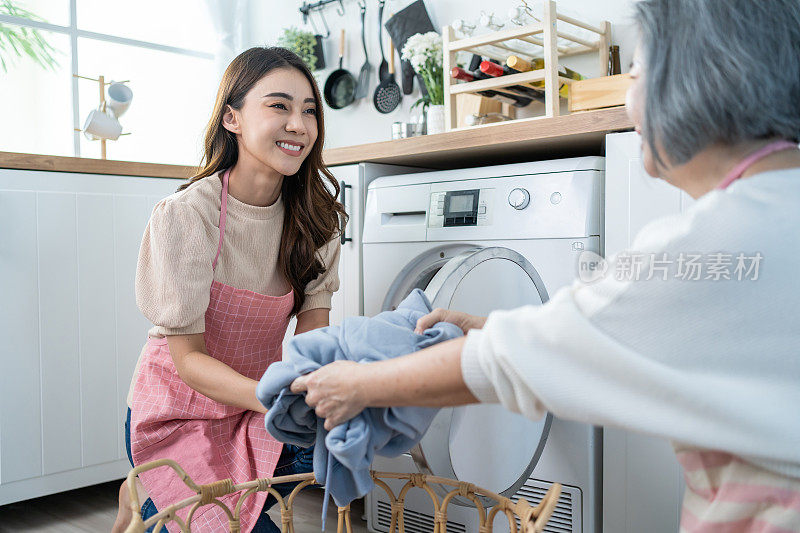 亚洲可爱的家庭在厨房里做家务和杂务。小女孩的女儿帮助和支持老母亲把衣服放进洗衣机里洗。清洁清洁的理念