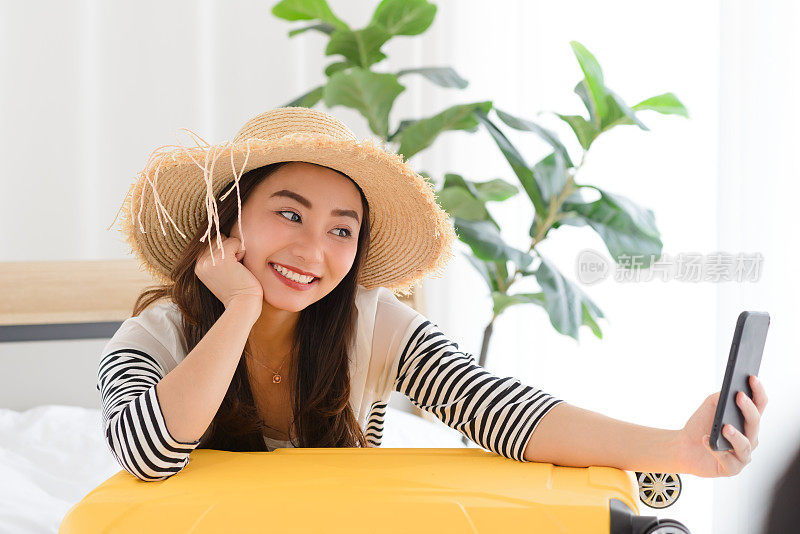 年轻的亚洲女性博主微笑在夏季休闲服装和草帽与facetime视频通话与智能手机在卧室。