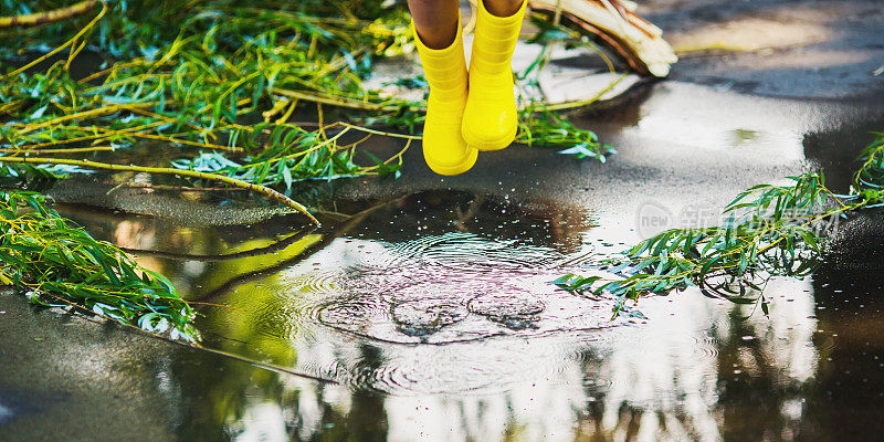 夏雨。水坑在沥青。穿着黄色雨靴的孩子在水坑里跳跃。孩子走过水坑。寒冷的秋天的雨。潮湿多雨的天气。水飞溅。水面全景图中的反射