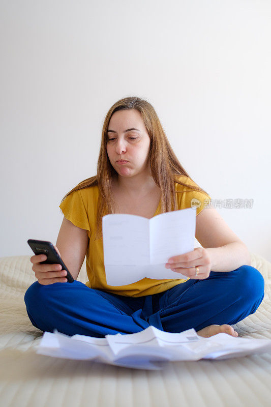 女人检查她的账单她的账单在她的床上，用手机计算器做数学，变得担心