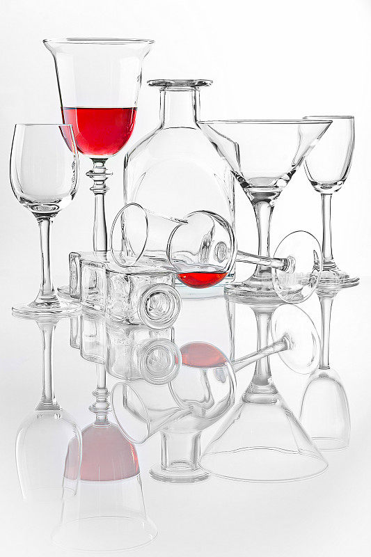 概念静物与不同的瓶子，酒杯，烈酒杯，马提尼杯和一些红色液体。工作室拍摄与充分的自然反射在白色背景。