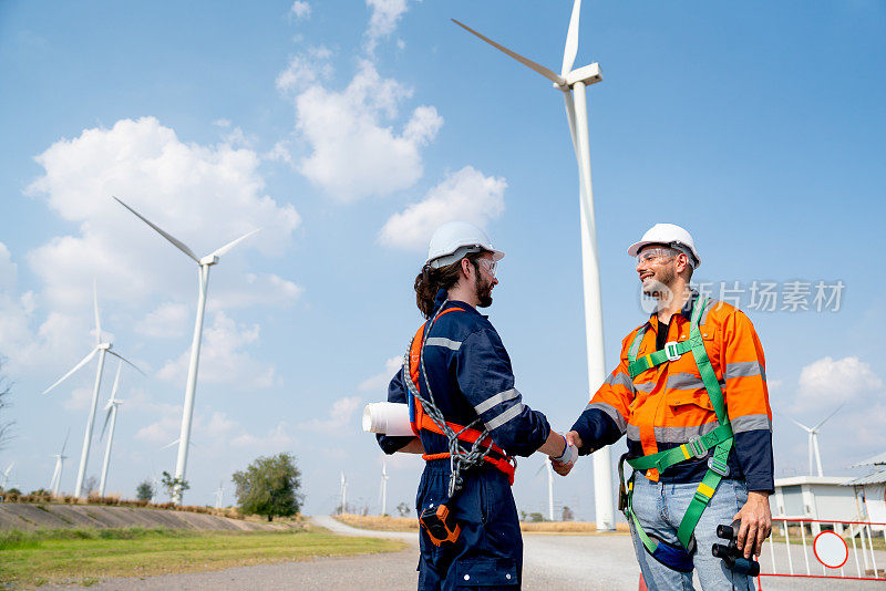 两名白人工程师或技术人员在工作场所项目成功后，在风车或风力涡轮机集群前握手。