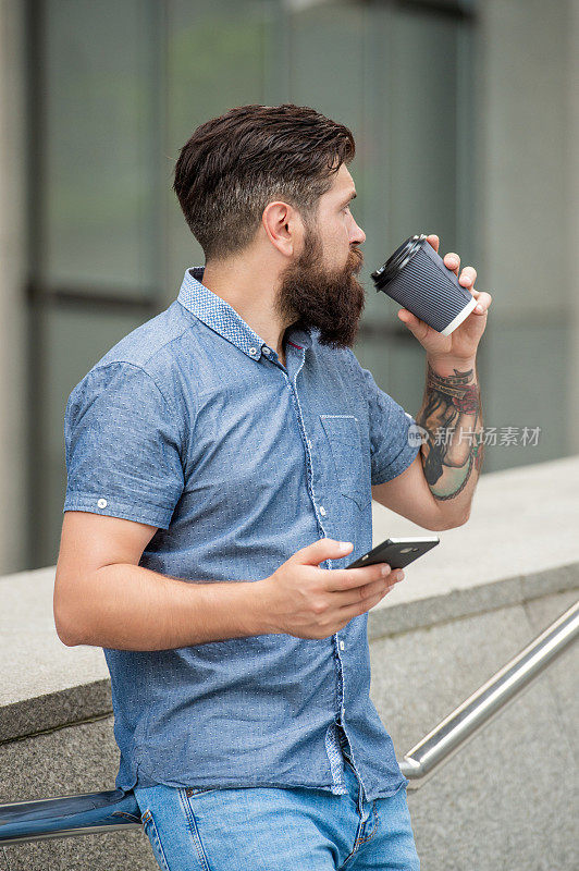 大胡子小伙一边在户外喝咖啡一边用手机发短信