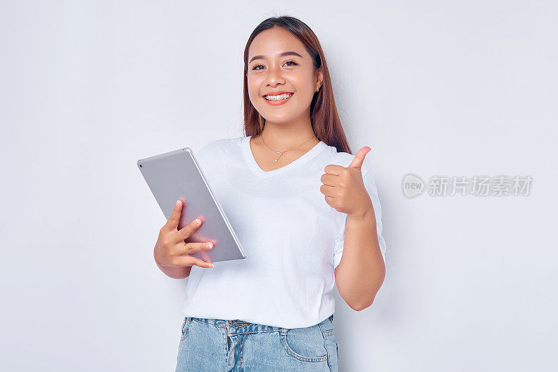 肖像微笑年轻的亚洲女孩在白色t恤拿着数字平板电脑，显示大拇指孤立在白色背景