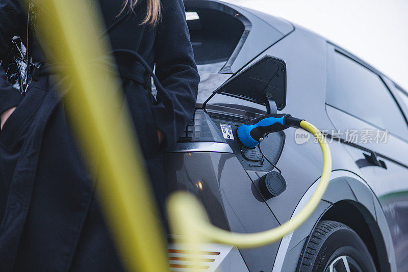 一辆电动汽车正在充电，一个穿大衣的女人站在旁边