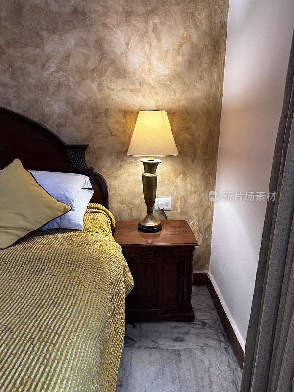 双人床旁的床头柜上照明的电灯图像，木质床头板，白色枕头与金色靠垫，金色床罩，重点在前景