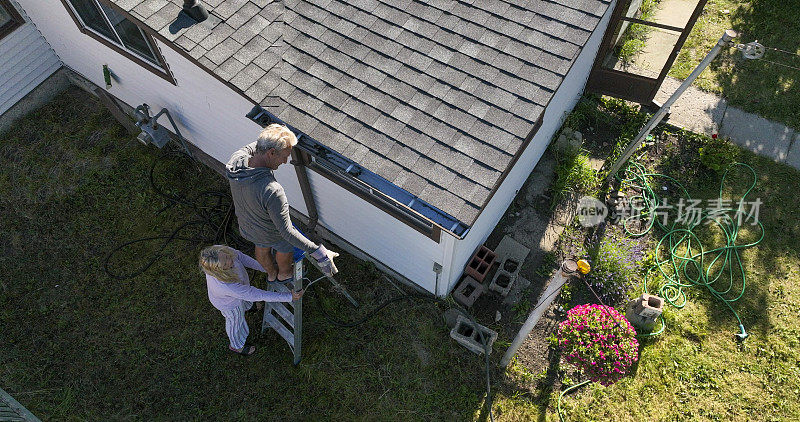 老人清理屋顶排水沟，他的同伴帮他扶梯子
