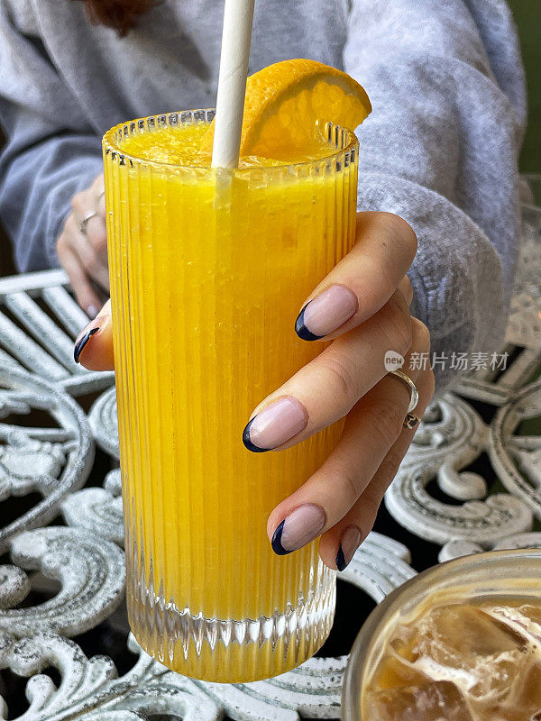 特写照片是一个不知名的女人拿着一杯橙汁和橙片，一杯冰咖啡放在餐厅的露天餐桌上，重点放在前景上