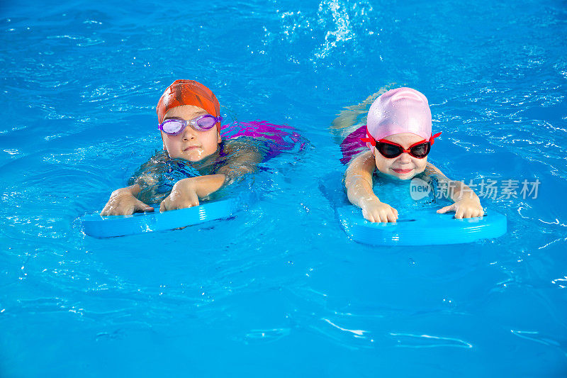 两个小女孩在泳池里玩得很开心，学习如何使用冲浪板游泳