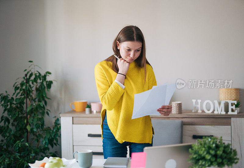 客厅里，一位年轻女子手里拿着一大堆文件，站在一台笔记本电脑前