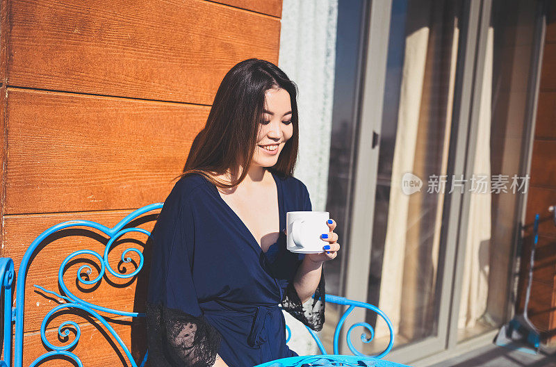 美丽的亚洲微笑女人享受咖啡或茶在阳光明媚的阳台。年轻快乐的女士穿着蓝色浴袍，夏天在露台上晒日光浴。