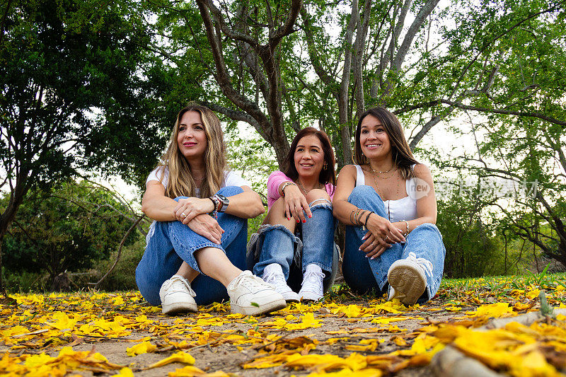 三位拉丁裔女性朋友坐在黄花丛中，带着惊奇或印象的表情望着远处的东西