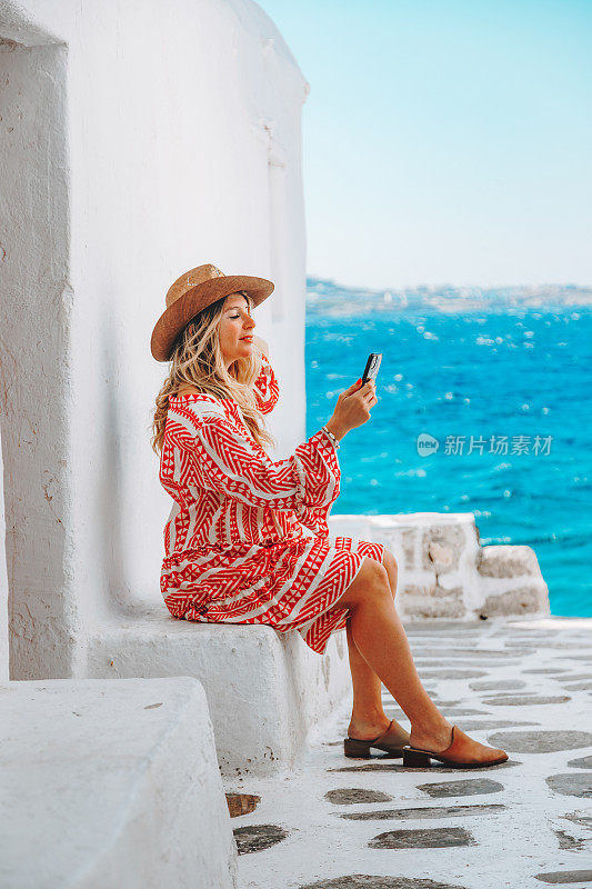 在一个阳光明媚的夏日，在米科诺斯岛一条狭窄的街道上，一位年轻的女游客微笑着看着她的智能手机，分享着她的内容，享受着爱琴海碧绿的海水。