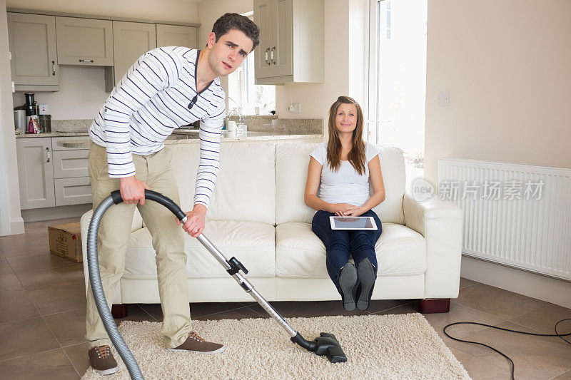 男人用吸尘器打扫地毯，而妻子正在沙发上休息