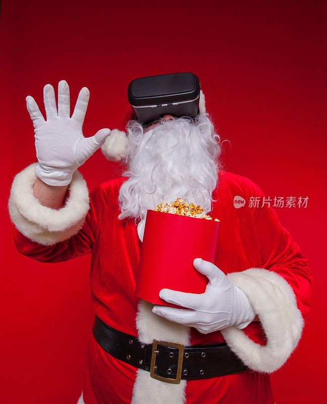 圣诞老人戴着虚拟现实眼镜和一个装满爆米花的红桶，在红色的背景上。圣诞节