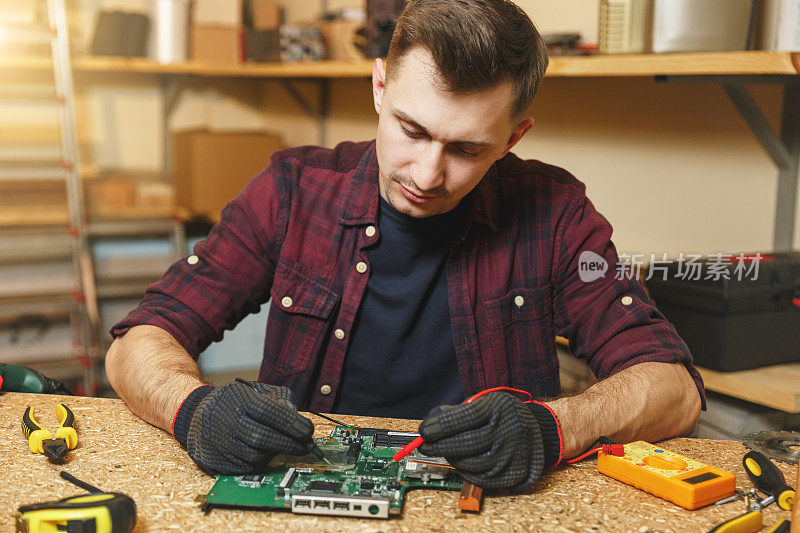对电工万用表。白人青年男子在格子衬衫，黑色t恤数字电子工程师修理，焊接电脑PC主板在车间的木桌与不同的工具。