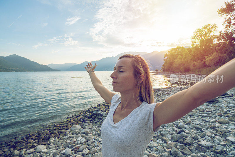 夕阳西下，拥抱大自然的快乐年轻女子;女性站在湖岸，以高山为背景——瑞士