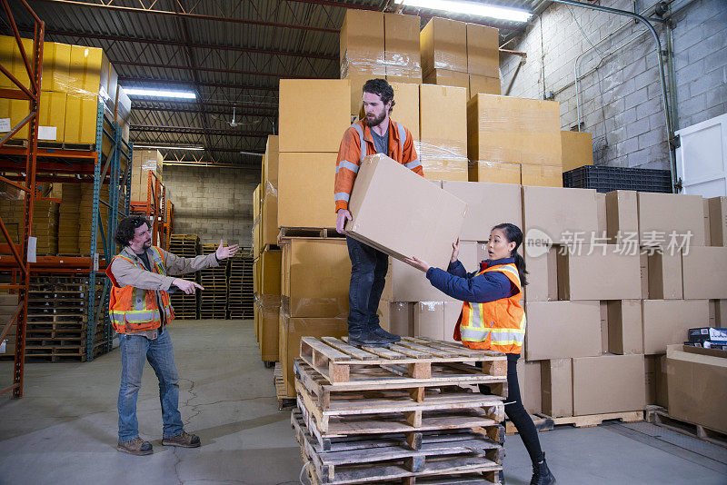 一个关于工业仓库工作场所安全的话题。一名员工不安全地站在一堆托盘上去够放置得更高的箱子。