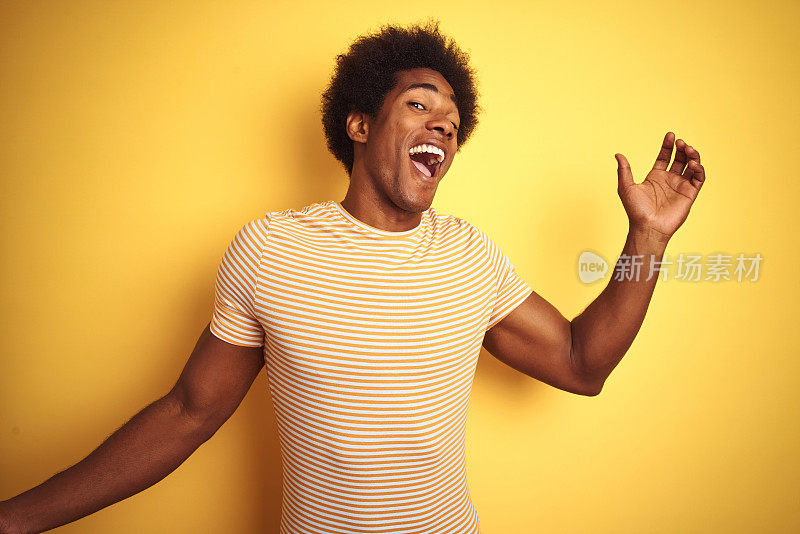 一个留着非洲式发型穿着条纹t恤的美国人站在孤立的黄色背景上，欢快地跳舞，微笑着听着音乐，随意而自信