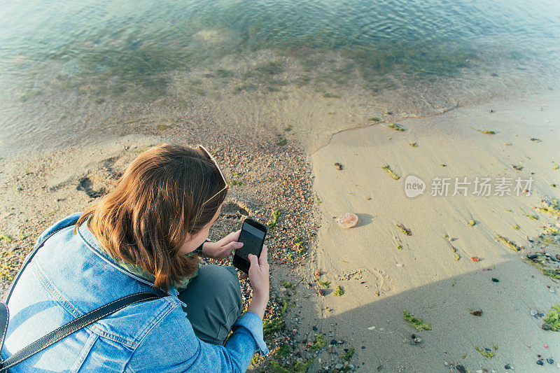 年轻的独自旅行者在海滩上用智能手机拍水母