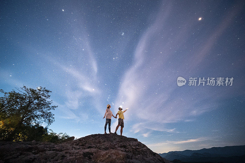 一起去摘星星。宽阔的镜头一对相爱的游客站在一个高岩石在晚上，看着宏伟的星空。灵感和灵性。心理健康。