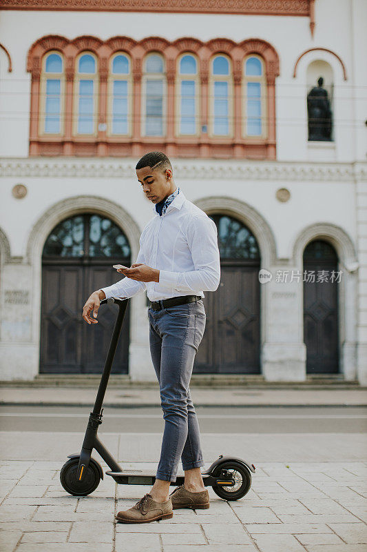 年轻的非洲裔美国人骑着电动车站在街上使用手机
