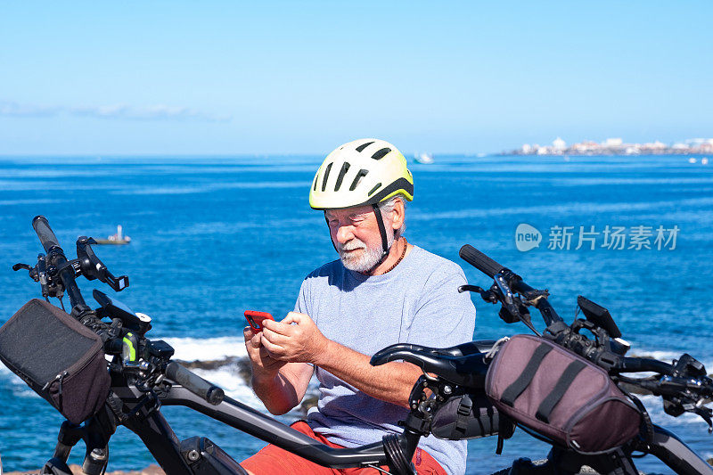 活跃的长者在户外远足时坐着并使用手机。他戴着自行车头盔靠近自行车，地平线在海上。积极的退休人员使用技术