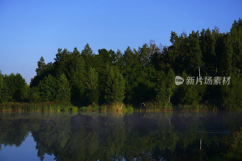 一个寒冷的夏日早晨，森林里有一个湖泊，森林的倒影和水面上的雾。雾和初升太阳的迷人风景。