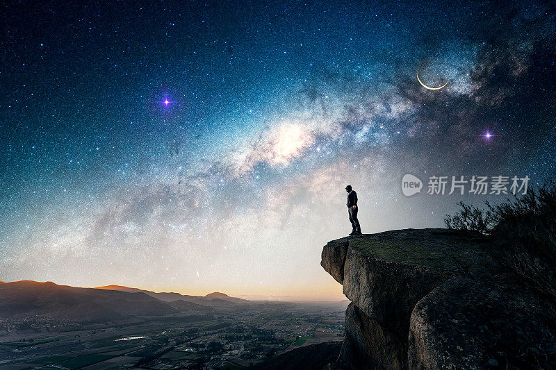 一个人站在户外的岩石上，越过山谷，凝视着银河和流星