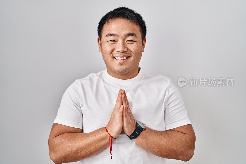 年轻的中国男子站在白色背景祈祷双手在一起请求原谅微笑自信。