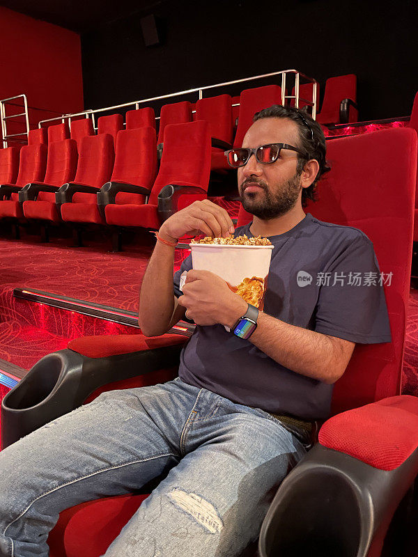 印度男子戴着3D眼镜在电影院看电影的特写图像，吃着一桶爆米花，一排排有饮料架的分层红色座位，靠过道的座位，重点放在前景