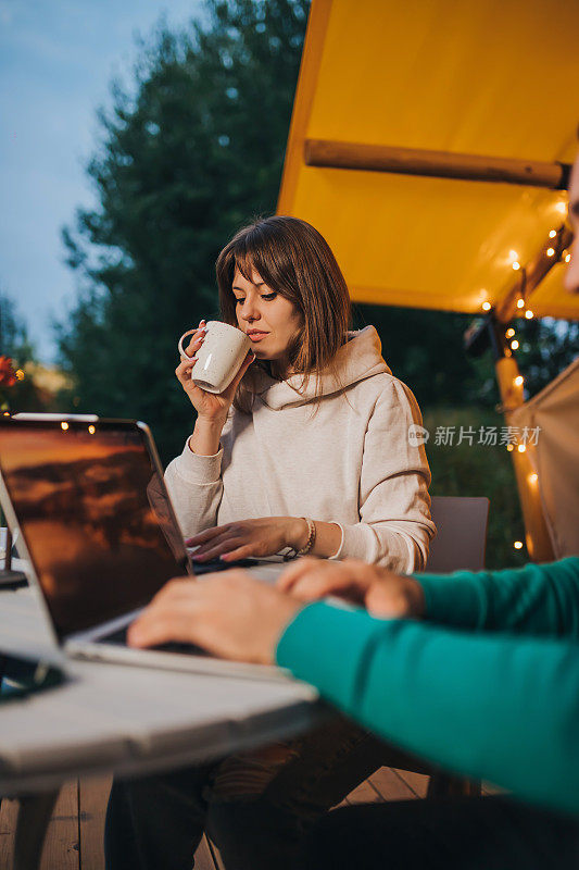 夏天的晚上，在舒适的野营帐篷里，女自由职业者在同事旁边喝着咖啡，用着笔记本电脑。用于户外度假和度假的豪华露营帐篷。生活方式的概念