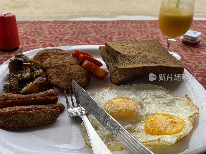 海滩餐厅的户外早餐场景特写图像，炸香肠，煎鸡蛋，番茄片，哈希饼，白盘子上的全麦吐司，单份黄油和果酱，刀，叉，一杯橙汁，重点在前景