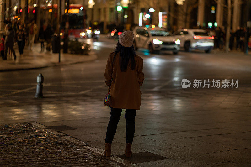 年轻女子的背影看着夜晚的城市灯光，穿着棕色的夹克