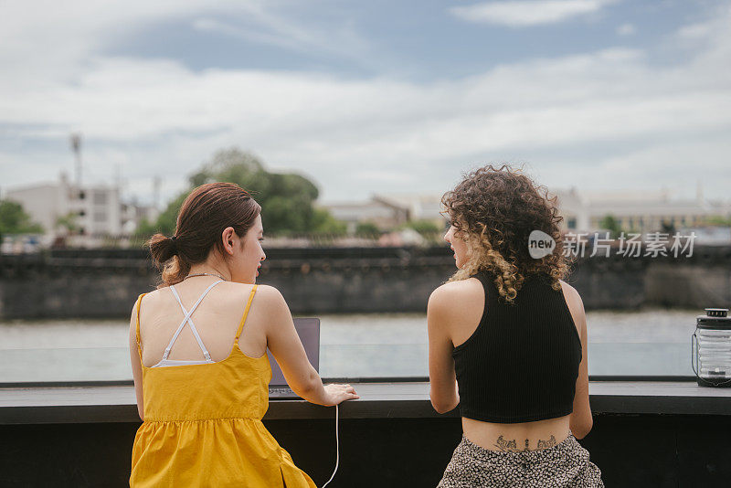 两个女人坐在咖啡馆里的后景，可以看到河边的美景。度假时一起聊天放松。