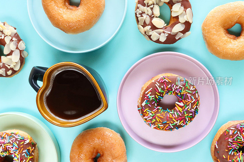 色彩柔和的盘子上有光滑的环形甜甜圈，用彩色糖屑或杏仁片装饰的普通和巧克力油炸面团甜点，心形黑咖啡杯，绿松石背景，高架视图